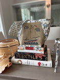 Load image into Gallery viewer, A handcrafted Nantucket quilted linen fabric tote bag with a Zweigart Penelope sailboat with waves and clouds front and back panel and floral hydrangea and lightship basket custom made linen fabric textile from our one of a kind artwork on the side panels that is quilted by our artisans. Handbag is styled on top of books on a wooden table next to a Erica Wilson needlepoint Nantucket lightship basket.