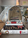 Load image into Gallery viewer, A custom artisan handcrafted self finishing needlepoint handbag created with quilted custom one of a kind artisan lightship baskets full of blue blooming New England hydrangeas and Nantucket cranberry fabric on the side panels. The front and back panels have Zweigart Penelope canvas with a sailboat, waves, clouds and flags inspired by Nantucket sailboats. Needlepoint canvas inset with a sailboat design, resting on top of a stack of books on a wooden table in a cozy indoor setting.